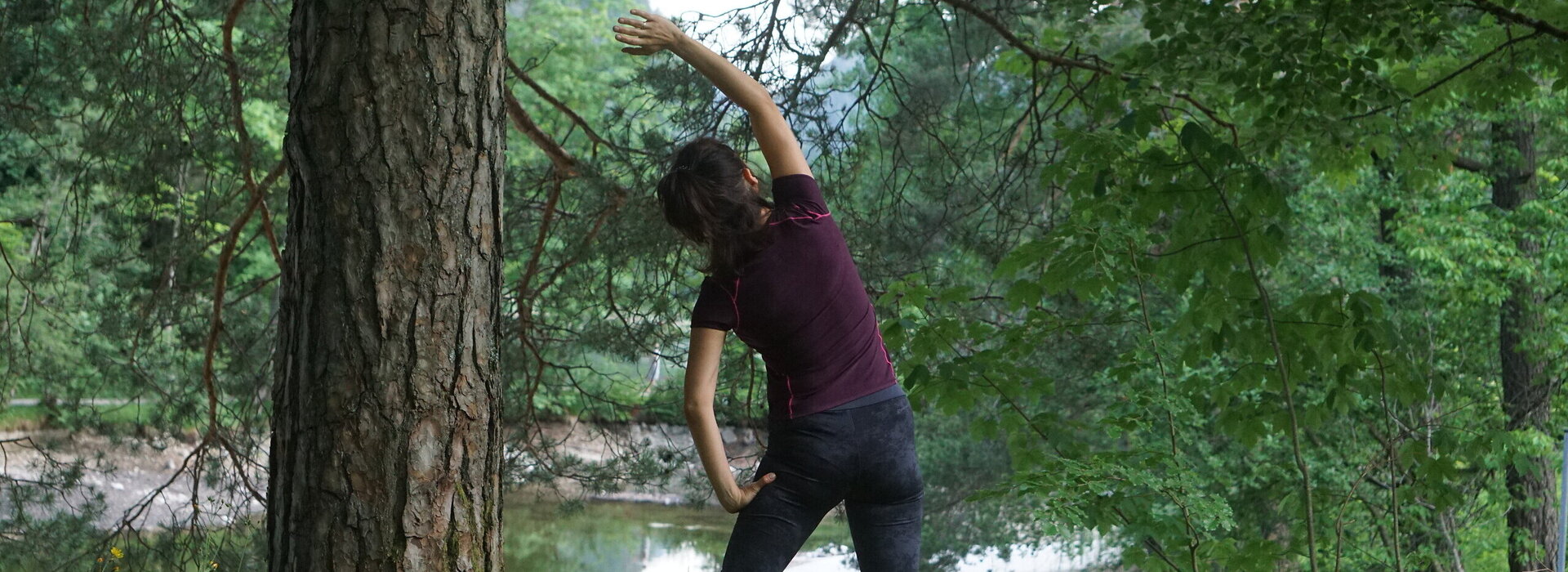 Das Bild zeigt eine Frau in sportlichem Outfit mit dunklen Haaren, die eine Yoga-Bewegung macht. Sie steht in der Natur unter einem Baum im hohen Gras und blickt auf ein Gewässer.
