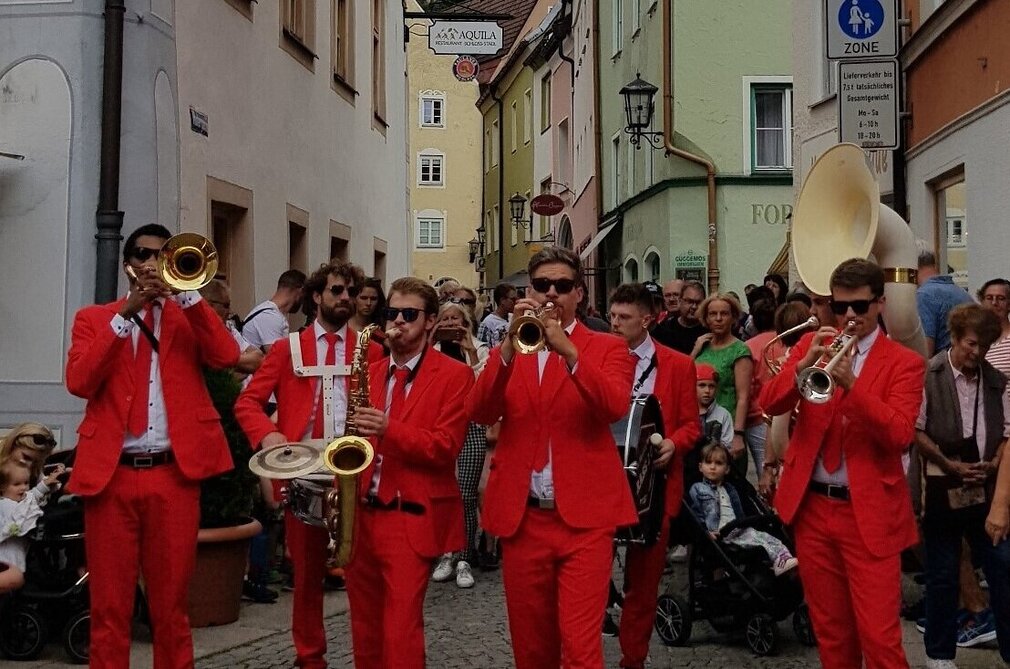 Man blickt in eine Gasse der Stadt Füssen. Im Vordergrund steht eine Bläsergruppe in roten Anzügen und weißen Hemden. Sie spielen Trompete, Posaune, Sousaphon, Saxophon und Schlafzeug. Hinter der Musikergruppe sind zahlreiche Menschen zu sehen. Der Himmel ist wolkenverhangen. 