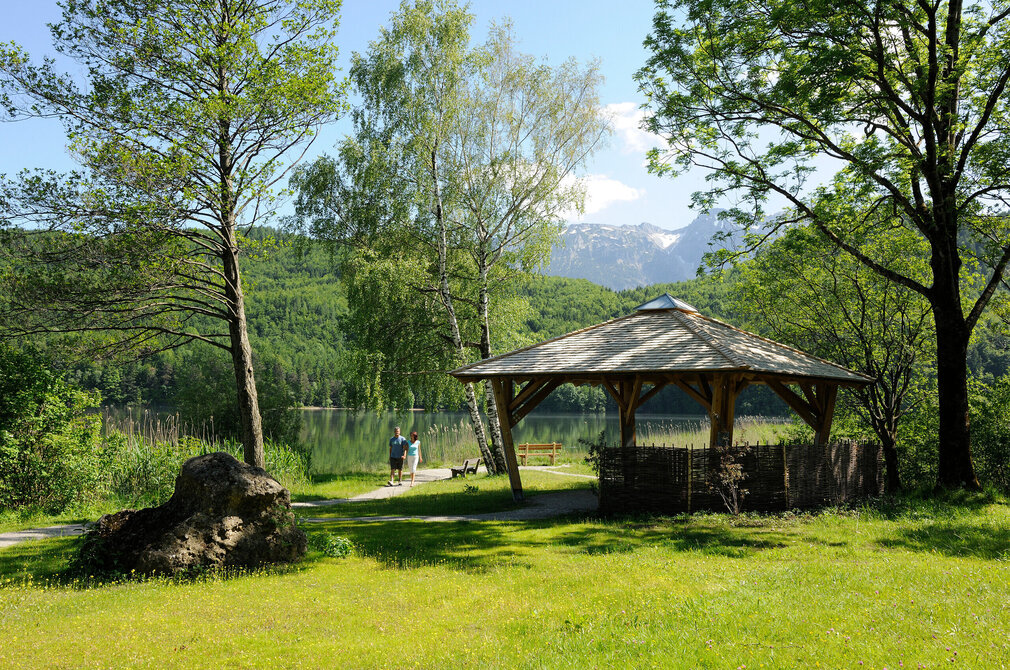 Das Bild zeigt zwei Personen, die auf einem Wanderweg laufen. Sie laufen vom Ufer eines Sees weg, vorbei an einem Holzpavillon. Vor ihnen ist eine Wiese zu sehen. Ringsum wachsen hohe Bäume. Im Hintergrund ist das andere Ufer des Sees zu sehen. Hier erheben sich bewaldete Hügel und Berge. Der Himmel ist blau. Die Sonne scheint. 