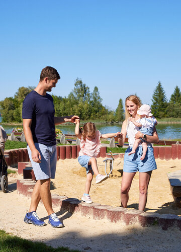 ie Szenene in dem Bild ist eine friedliche Momentaufnahme in einem Park. Vor einem ruhigen See stehen ein Mann und eine Frau, beide tragen legeres Outfit. Der Mann hat ein blaues T-Shirt und blaue Shorts an, während die Frau ein weißes T-Shirt und blaue Shorts trägt. Sie halten Händchen, was ihre Nähe zum Ausdruck bringt.  Im Hintergrund ist ein friedlicher Teich zu sehen, das zur friedlichen Atmosphäre des Parks beiträgt. Einige Bäume stehen dort und bieten eine natürliche Kulisse für die Szene.  Der Park ist auch mit einem Spielplatz ausgestattet, inklusive eines Sandkastens und eines Schaukelsatzes. Der Sandkasten liegt nahe zum See, bietet also eine fröhliche und aktive Spielmöglichkeit für Kinder. Der Schaukelsatz befindet sich etwas weiter weg, wo Kinder ungetehrt spielen und Eltern ihnen zuschauen können.  Das Bild zeigt generell einen typischen Tag im Park, mit Leuten, die die Natur genießen, und Kindern, die im Sandkasten und auf den Schaukeln spielen.
