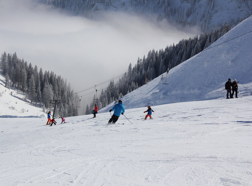 Das Bild zeigt eine abfallende Skipiste auf der mehrere Personen, auch Kinder Skifahren. Neben der Piste läuft der Schlepplift. Im Tal liegt eine dicke Nebelschicht. Die Landschaft ringsum ist tief verschneit. Die Sonne scheint.  