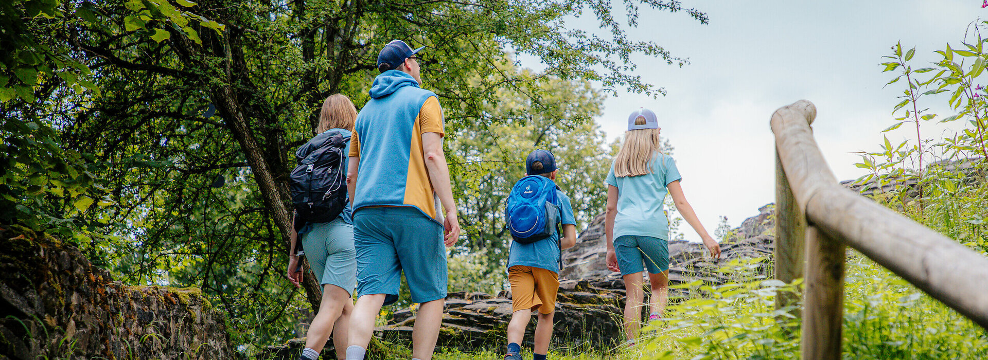 Das Bild zeigt eine Familie, bestehend aus einer Frau, einem Mann, einem Jungen und einem Mädchen, die einen Wanderweg nach oben gehen. Sie tragen sportliche Freizeitkleidung. Im Hintergrund ist die Mauer einer Ruine zu sehen. Ringsum wachsen Bäume, Büsche und Gräser. Der Himmel ist blau. Die Sonne scheint.