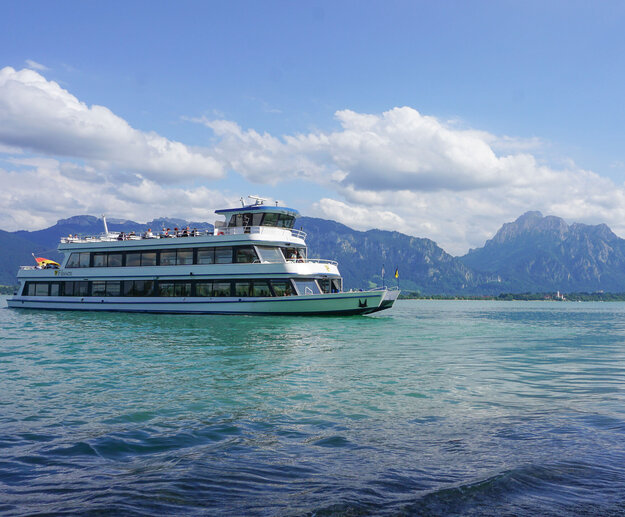 Das Bild fängt eine friedliche Szene eines weißen Schiffes mit drei Aussichtsdecks ein, das auf einem ruhigen blauen See – dem Forggensee - schwimmt. Das Schiff steht im Mittelpunkt des Bildes und wird von der unendlichen Weite des Forggensees umgeben. Der See selbst ist inmitten einer malerischen Kulisse von majestätischen Bergen eingebettet. Der klare und blaue Himmel ist vereinzelt von flauschigen, weißen Wolken durchzogen. 