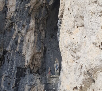 Das Bild zeigt einen atemberaubenden Blick auf eine grotteähnliche Struktur, möglicherweise ein Tempel oder ein Heiligtum, die in eine felsige Klippe eingebettet ist. Der Eingang zur Grotte ist ein kleiner Bogen, der Besucher einlädt, die Tiefe zu erkunden. In der Grotte kann eine Statue einer Person gesehen werden, was der Szene einen Hauch von Spiritualität hinzufügt. Die Grotte wird von einer üppigen grünen Landschaft umgeben, mit einem kleinen Baum im Vordergrund sichtbar. Die Perspektive des Bildes ist von einem niedrigen Winkel, der auf die Grotte blickt, was die Größe der Struktur unterstreicht. Die Farben in dem Bild sind hauptsächlich erdige Töne, wobei das Grün der Landschaft wunderschön mit dem Grau der felsigen Klippe kontrastiert. Das Bild ist ein Zeugnis der Schönheit der Natur und die Faszination menschgemachter Strukturen.