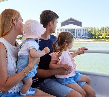 Das Bild zeigt einen friedlichen Moment auf einem Boot. Eine Familie von vier Personen sitzt zusammen und genießt die Aussicht auf das Wasser. Die Mutter, in einem weißen Bluse gekleidet, hält ihr Baby auf dem Arm. Der Vater trägt ein blaues Hemd und hält ihre Tochter auf dem Schoß. Die Tochter ist in einem rosa Shirt gekleidet und sitzt auf dem Schoß des Vaters. Das Boot, auf dem sie sind, ist weiß und verfügt über eine Reling. Im Hintergrund ist ein Gebäude und Bäume zu sehen, die zur malerischen Szene beitragen. Die Familie scheint einen großartigen Tag zusammen zu verbringen und genießt ihre Bootsfahrt voll und ganz.