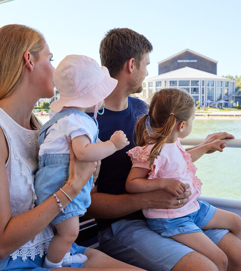 Das Bild zeigt einen friedlichen Moment auf einem Boot. Eine Familie von vier Personen sitzt zusammen und genießt die Aussicht auf das Wasser. Die Mutter, in einem weißen Bluse gekleidet, hält ihr Baby auf dem Arm. Der Vater trägt ein blaues Hemd und hält ihre Tochter auf dem Schoß. Die Tochter ist in einem rosa Shirt gekleidet und sitzt auf dem Schoß des Vaters. Das Boot, auf dem sie sind, ist weiß und verfügt über eine Reling. Im Hintergrund ist ein Gebäude und Bäume zu sehen, die zur malerischen Szene beitragen. Die Familie scheint einen großartigen Tag zusammen zu verbringen und genießt ihre Bootsfahrt voll und ganz.