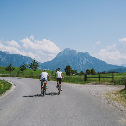 Das Bild zeigt einen Mann und eine Frau beim Fahrradfahren. Sie tragen Freizeitkleidung und Helme und fahren vom Betrachter weg auf einer asphaltierten Straße. Die Straße ist von Wiesen umgeben. Im Hintergrund erheben sich die Berge. Die Sonne scheint. Der Himmel ist blau. 