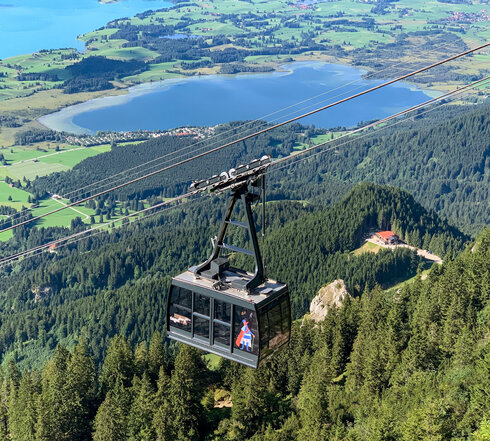 Das Bild zeigt die an einem Seil hängende Kabine einer Bergbahn. Im Tal erblickt man zwei blaue Seen, Wiesen und Wälder. Die Sonne scheint. 
