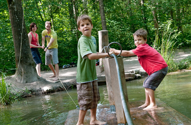 Das Bild zeigt zwei Jungen, die auf einem hölzernen Floß über einen Bach fahren. Dabei halten sie sich an einem Stahlseil fest, das über den Bach gespannt ist. Im Hintergrund stehen ein Mann und eine Frau und lenken das Floß. Ringsum wachsen hohe Bäume. Die Sonne scheint. Das Wasser des Bachs ist trüb. Die Kinder haben sichtlich Spaß.