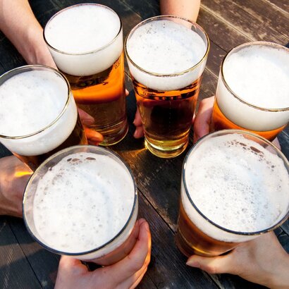 hier erkennt man fünf Hände, die mit vollen Biergläsern die auf einem Holztisch  anstoßen. 