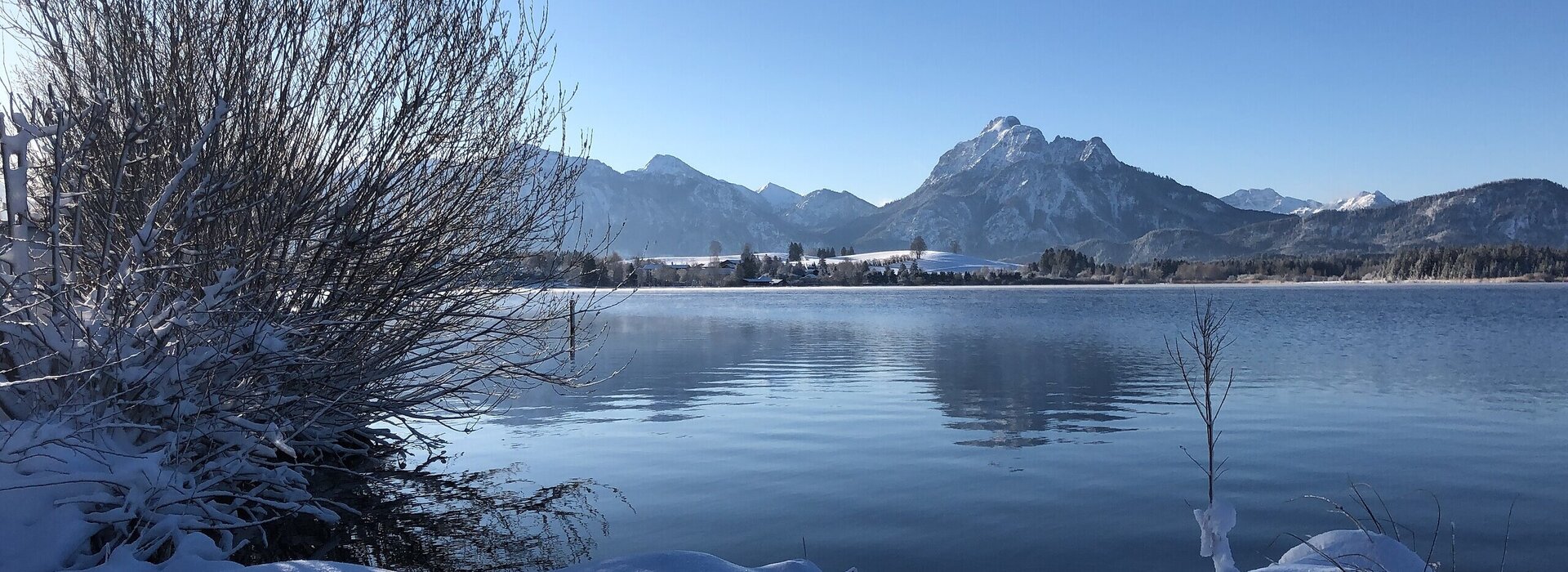 Das Bild zeigt eine verschneite Winterszene. Im Fokus steht ein See mit ruhiger Wasseroberfläche. Im Hintergrund erheben sich die schneebedeckten Berge. Der Himmel ist blau. Die Sonne scheint. 
