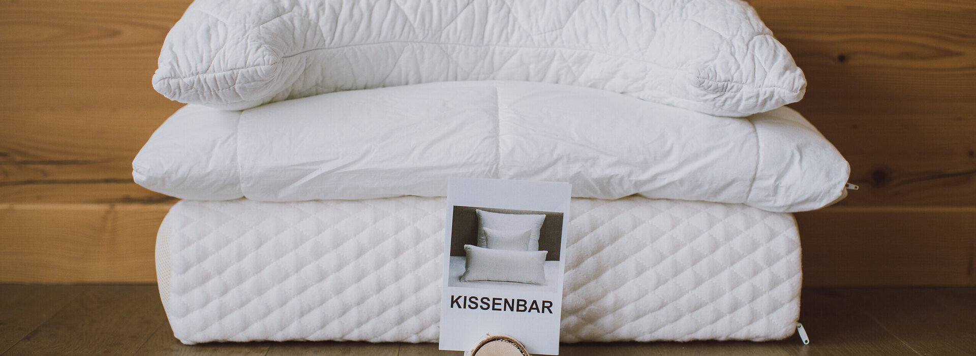 Das Bild zeigt drei unterschiedliche, aufeinanderliegende Kopfkissen, ohne Bezug, vor einer Holzwand. Auf dem obersten Kissen steht „Relax“ in Holzbuchstaben. Vor dem Kissenstapel steht ein Papierschild mit der Aufschrift „Kissenstapel“ in einer Holzhalterung. 