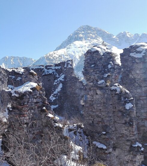 Das Bild zeigt die Mauerreste einer Burg. Es ist Winter. Die Mauerreste sind schneebedeckt.  Im Hintergrund erheben sich schneebedeckte Berge. Der Himmel ist blau. Die Sonne scheint. 