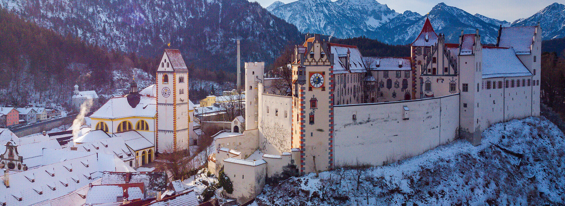 Das Bild fängt die atemberaubende Sicht auf das mittelalterliche Städtchen Füssen in Deutschland ein. Die Stadt liegt auf einem Hügel, von majestätischen Bergen umgeben. Die Architektur der Stadt ist durch bunte Gebäude geprägt, jedes mit einem einzigartigen Charme. Die Gebäude sind mit Schnee verziert, was eine Prise Winter in die Szene bringt