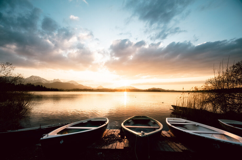 Das Bild zeigt einen Sonnenuntergang am Ufer des Hopfensees. Der Himmel ist in orange und blau gefärbt und dient als beeindruckende Kulisse für den ruhigen See. Am Ufer des Sees liegen, im Zentrum des Bildes drei Ruderboote an Land. Die Ruderboote, die in Größe und Verarbeitung unterschiedlich sind, sind leer und warten still auf ihr nächstes Abenteuer. 