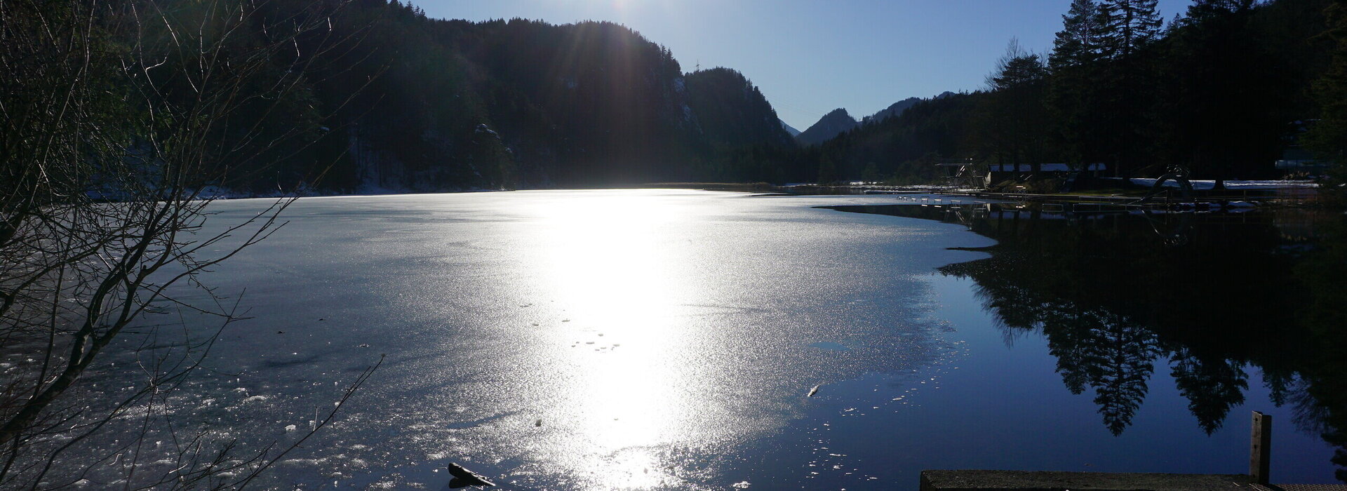 Man blickt auf einen teilweise zugefrorenen See, der von Hügeln und Wäldern umrahmt ist. Das Ufer ist schneebedeckt. Der Himmel ist blau. Die Sonne scheint.  
