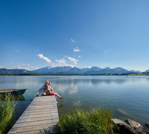 Auf dem Bild ist eine idyllische Szene am Hopfensee zu sehen. Es ist Sommer. Eine Frau kniet hinter einem Mann auf einem Holzsteg und umarmt ihn von hinten. Er sitzt auf dem Holzsteg. Seine Beine baumeln über dem Rand des Stegs herunter. Seine Füße berühren leicht die Wasseroberfläche des tiefblauen Sees. Beide blicken in die Ferne und lächeln. Der Holzsteg ragt in das ruhige Wasser des Hopfensees hinein, welcher von Bergen umgeben ist. Der Himmel ist blau und nur leicht durchzogen von wenigen Wolken. In der Ferne sind ein paar Ruder- und Tretboote auf dem Wasser zu sehen.