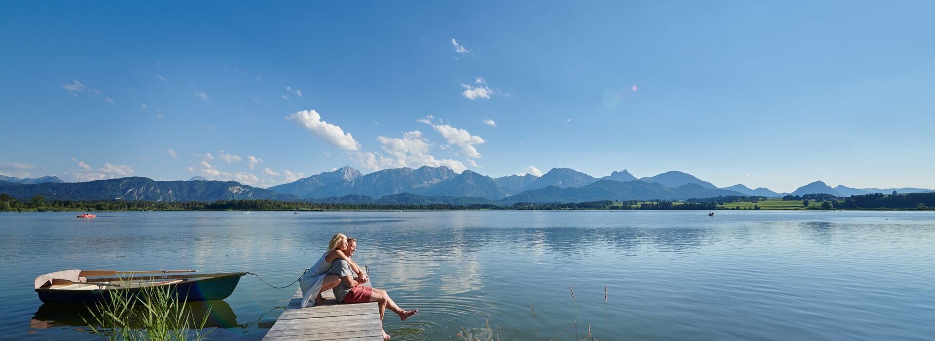 Auf dem Bild ist eine idyllische Szene am Hopfensee zu sehen. Es ist Sommer. Eine Frau kniet hinter einem Mann auf einem Holzsteg und umarmt ihn von hinten. Er sitzt auf dem Holzsteg. Seine Beine baumeln über dem Rand des Stegs herunter. Seine Füße berühren leicht die Wasseroberfläche des tiefblauen Sees. Beide blicken in die Ferne und lächeln. Der Holzsteg ragt in das ruhige Wasser des Hopfensees hinein, welcher von Bergen umgeben ist. Der Himmel ist blau und nur leicht durchzogen von wenigen Wolken. In der Ferne sind ein paar Ruder- und Tretboote auf dem Wasser zu sehen.