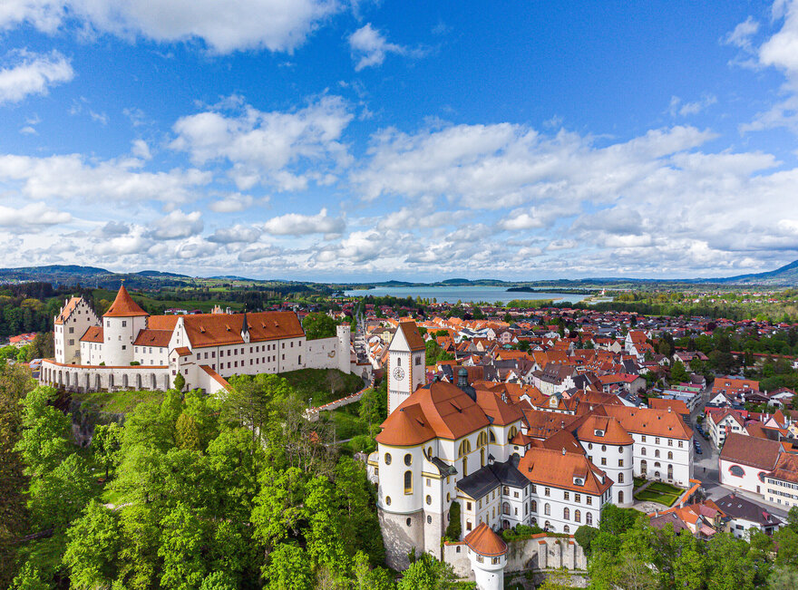 Die Abbildung zeigt eine malerische Stadt mit einer Burgenstruktur, dem Hohen Schloss und auf das alte Kloster St. Mang, die von reichlich grünen Bäumen umgeben ist. Die Stadt ist in einem Tal gelegen, mit einem Fluss, der durch sie hindurchfließt. Im Hintergrund sieht man den Forggensee.