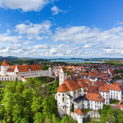 Die Abbildung zeigt eine malerische Stadt mit einer Burgenstruktur, dem Hohen Schloss und auf das alte Kloster St. Mang, die von reichlich grünen Bäumen umgeben ist. Die Stadt ist in einem Tal gelegen, mit einem Fluss, der durch sie hindurchfließt. Im Hintergrund sieht man den Forggensee.