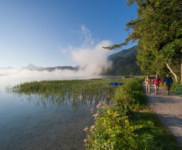 Das Bild zeigt drei Personen in Wanderkleidung und mit Rucksäcken, die am Ufer eine Sees stehen. Sie wandern am Ufer entlang. Über dem Wasser wabern Nebelschwaden. Die Sonne scheint der Himmel ist blau. Im Hintergrund sieht man hohe Berge und bewaldete Hügel.     
