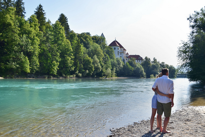 Das Bild zeigt ein Paar, das an einem steinigen Ufer des Flusses Lech steht. Der Mann und die Frau stehen dicht beieinander, ihre Körper sind dem Wasser zugewandt. Der türkise Fluss liegt im Fokus des Bildes. Auf der gegenüberliegenden Seite erhebt sich ein großer Gebäudekomplex mit weißen Mauern und einem roten Dach. Hierbei handelt es sich um das Hohe Schloss und das ehemalige Kloster St. Mang in Füssen. Der Himmel darüber ist klar, blau und wolkenlos.