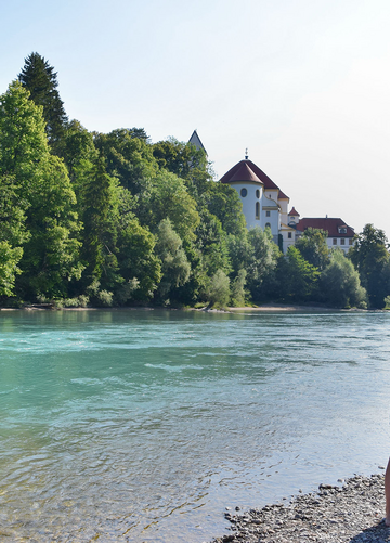 Das Bild zeigt ein Paar, das an einem steinigen Ufer des Flusses Lech steht. Der Mann und die Frau stehen dicht beieinander, ihre Körper sind dem Wasser zugewandt. Der türkise Fluss liegt im Fokus des Bildes. Auf der gegenüberliegenden Seite erhebt sich ein großer Gebäudekomplex mit weißen Mauern und einem roten Dach. Hierbei handelt es sich um das Hohe Schloss und das ehemalige Kloster St. Mang in Füssen. Der Himmel darüber ist klar, blau und wolkenlos.