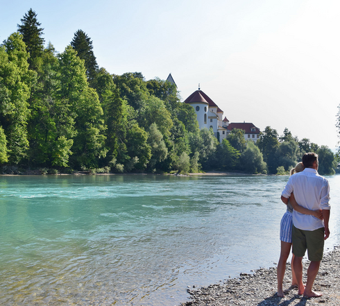 Das Bild zeigt ein Paar, das an einem steinigen Ufer des Flusses Lech steht. Der Mann und die Frau stehen dicht beieinander, ihre Körper sind dem Wasser zugewandt. Der türkise Fluss liegt im Fokus des Bildes. Auf der gegenüberliegenden Seite erhebt sich ein großer Gebäudekomplex mit weißen Mauern und einem roten Dach. Hierbei handelt es sich um das Hohe Schloss und das ehemalige Kloster St. Mang in Füssen. Der Himmel darüber ist klar, blau und wolkenlos.