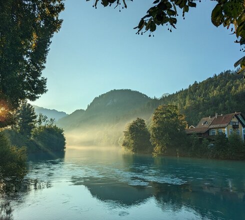 Das Bild zeigt eine ruhige Landschaft, die von einem friedlichen Fluss beherrscht wird, der sich durch die Szene schlängelt. Der türkise Fluss Lech fließt friedlich dahin, eingebettet in üppige Vegetation, mit Bäumen und Sträuchern, die die Ufer säumen. Die ruhige Oberfläche des Flusses spiegelt die umgebende Landschaft wider. Auf der rechten Flussseite steht ein charmantes gelbes Haus mit rotem Dach. Der klare und blaue Morgenhimmel ist wolkenlos. Die Sonne ist gerade aufgegangen. Ihre Strahlen bahnen sich ihren Weg durch die Äste der Bäume, die am Ufer stehen. 