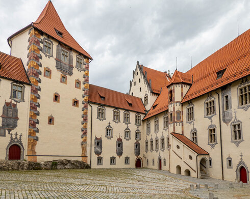 Das Bild zeigt ein große mit Illusionsmalerei verziertes Gebäude. Es hat viele Fenster und rote Dächer. Der Gebäudekomplexes besteht aus mehreren unterschiedlichen Gebäudeteilen, die aneinandergebaut sind. Der Himmel darüber ist wolkenverhangen.