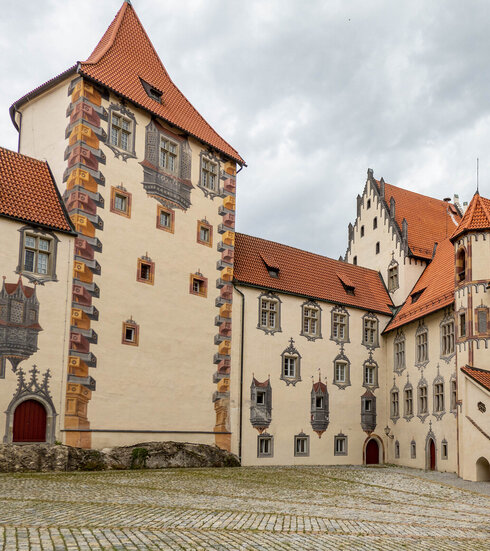 Das Bild zeigt ein große mit Illusionsmalerei verziertes Gebäude. Es hat viele Fenster und rote Dächer. Der Gebäudekomplexes besteht aus mehreren unterschiedlichen Gebäudeteilen, die aneinandergebaut sind. Der Himmel darüber ist wolkenverhangen.