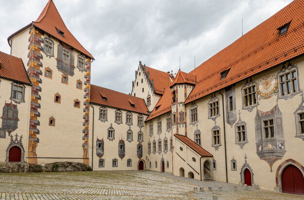 Das Bild zeigt ein große mit Illusionsmalerei verziertes Gebäude. Es hat viele Fenster und rote Dächer. Der Gebäudekomplexes besteht aus mehreren unterschiedlichen Gebäudeteilen, die aneinandergebaut sind. Der Himmel darüber ist wolkenverhangen.