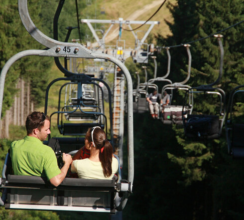 Das Bild zeigt eine Mann und eine Frau, die auf einem Zweisitzer einer Sesselbahn fahren. Auch in weiteren Zweisitzern sind Personen zu sehen. Links und rechts der Bahnwachsen hohe grüne Bäume. Die Sonne scheint.