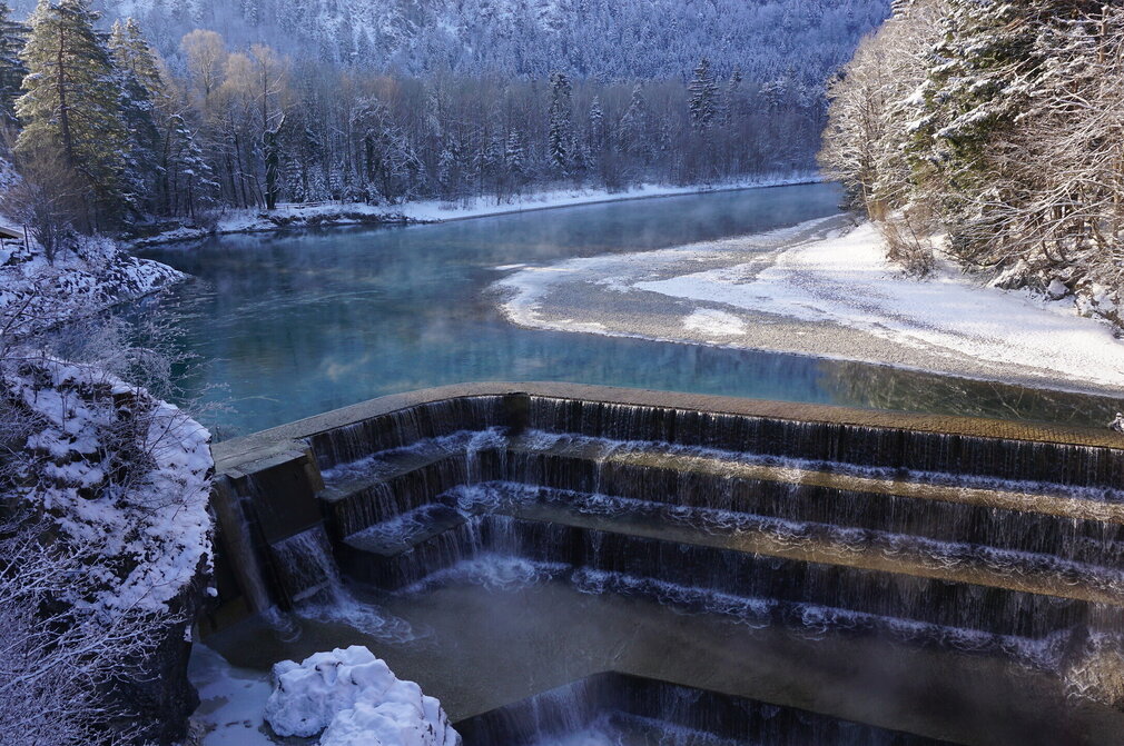 Das Bild zeigt eine friedliche Winterlandschaft. Ein dunkelblauer Fluss schlängelt sich durch die Landschaft. Der Fluss wird von einem schneebedeckten Wald flankiert, dessen Bäume sich gegen den Himmel abheben. Der Fluss fließt über gemauerte Steinstufen hinab. Der Himmel ist klar und blau, was einen wunderschönen Kontrast zum verschneiten Panorama bildet. Das Bild wird von einem hohen Standpunkt aus aufgenommen, der einen Panoramablick auf den Fluss und seine Umgebung bietet. 