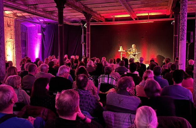 Das Bild zeigt eine Frau mit Gitarre, die auf einer Bühne sitzt. Vor ihr sitzen zahlreiche, ihr zugewandte Menschen auf Stühlen. Der Raum ist pink beleuchtet. 