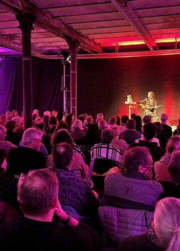 Das Bild zeigt eine Frau mit Gitarre, die auf einer Bühne sitzt. Vor ihr sitzen zahlreiche, ihr zugewandte Menschen auf Stühlen. Der Raum ist pink beleuchtet. 