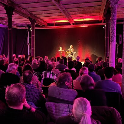 Das Bild zeigt eine Frau mit Gitarre, die auf einer Bühne sitzt. Vor ihr sitzen zahlreiche, ihr zugewandte Menschen auf Stühlen. Der Raum ist pink beleuchtet. 