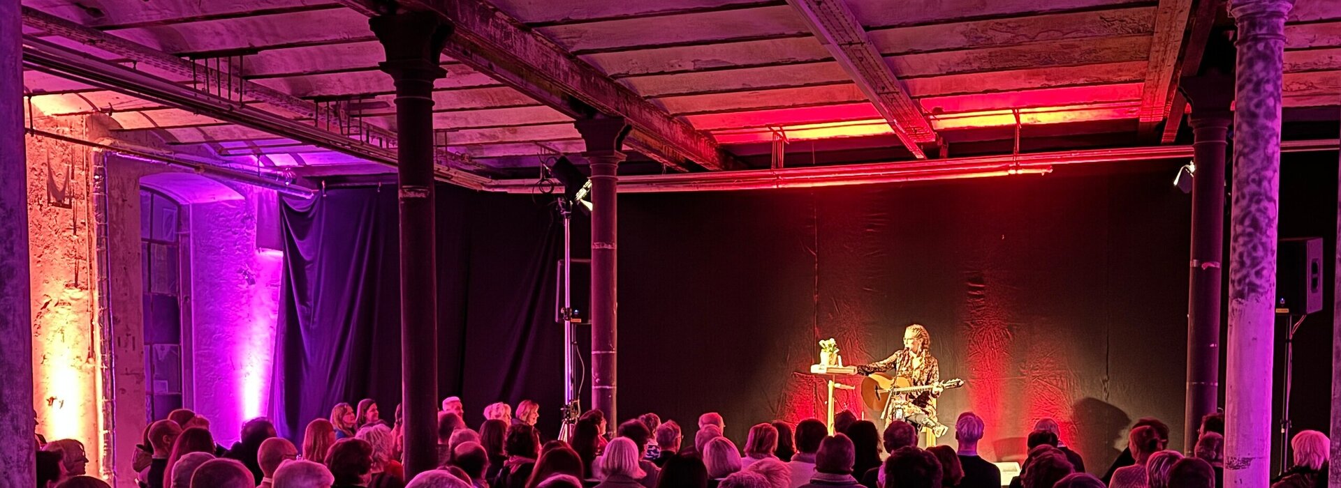 Das Bild zeigt eine Frau mit Gitarre, die auf einer Bühne sitzt. Vor ihr sitzen zahlreiche, ihr zugewandte Menschen auf Stühlen. Der Raum ist pink beleuchtet. 