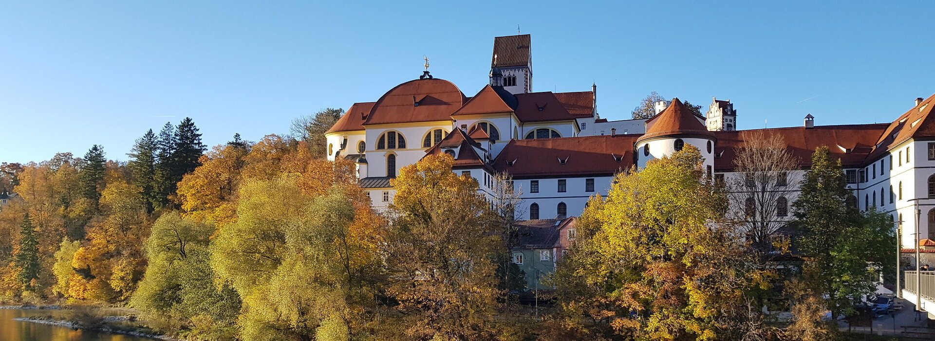 Das Bild zeigt den Blick auf das ehemalige Kloster St. Mang in Füssen im Allgäu vom gegenüberliegenden Lechufer aus. Der brocke Klosterbau erhebt sich über dem herbstlich gefärbten Baumbestand entlang des Flusses Lech. 