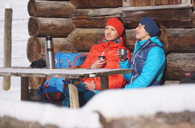 Das Bild zeigt zwei Frauen, die an einem Holztisch vor einem Holzstadel sitzen. Nur ein Detail der Wand ist zu sehen. Beide tragen Winterkleidung und Stirnbänder. Sie haben je einen Thermoskannenbecher in der Hand. Die Thermoskannen stehen auf dem Tisch. Die Sonne scheint. Ringsum ist alles schneebedeckt. 