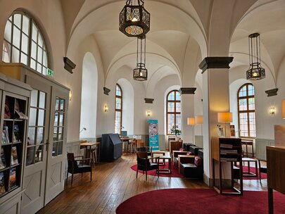 Man blickt in einen hohen Raum mit Kreuzrippengewölbe und Bogenfenster, der mit Säulen unterbrochen ist. Im Raum stehen mehrere unterschiedliche Sitzgelegenheiten verteilt. Schwarze Ledersessel mit Holzlehnen und niedrigen Holztischen stehe in der Mitte des Raumes. Vor den Fenstern sind Schreibtische zu sehen. Auf den Ledersesseln liegen rote Kissen. Auf dem Holzdielenboden liegen rote, runde Teppiche. Im Vordergrund sind Vitrinen mit Zeitschriften zu sehen. Der Raum ist hell und einladend.