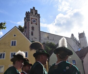 Das Bild zeigt mehrere Personen in Tracht. Man erkennt nur ihre Köpfe und Oberkörper. Die Männer tragen grüne Trachtenjacken. Die Frau mit Blüten verzierte Schultertücher. Alle tragen grüne Trachtenhüte. Die Männer haben Gamsbärte, die Frauen Adlerflaume an ihren Hüten befestigt. Im Hintergrund sind bunte Hausfassaden zu erkennen. Darüber thront der Uhrturm des Hohen Schlosses. Der Himmel ist blau. Die Sonne scheint.