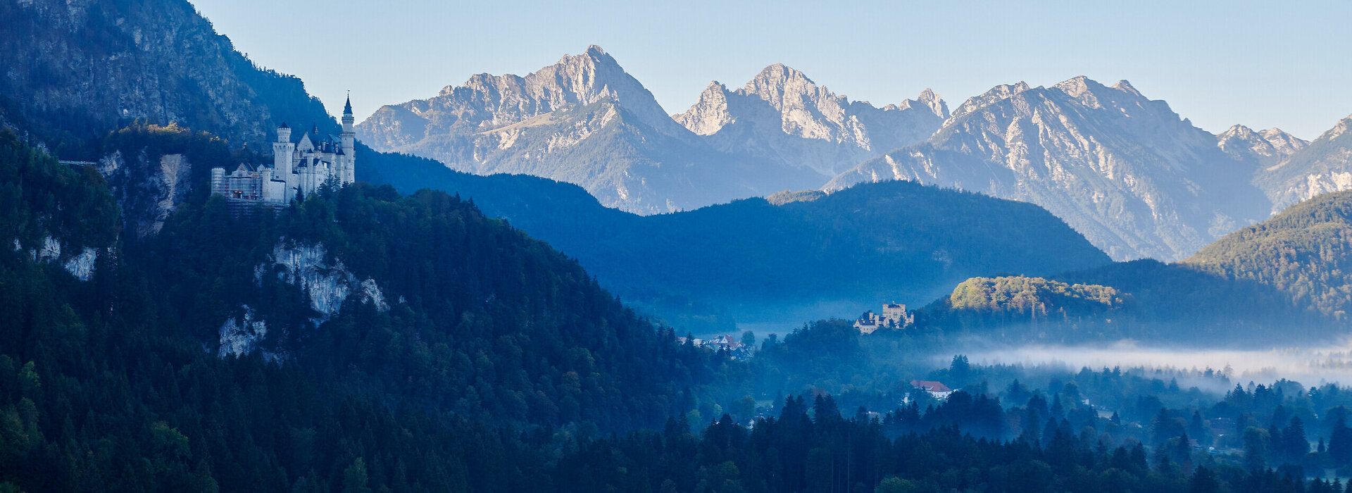 Das Bild zeigt die beiden Schlösser Hohenschwangau und Neuschwanstein, die auf zwei Anhöhen über den umliegenden Wiesen und Wäldern thronen. Im Hintergrund erheben sich die Berge. Der Himmel ist blau. Die Sonne geht gerade auf. Die Schlösser liegen noch im Schatten. 