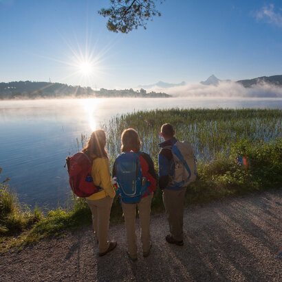 Das Bild zeigt drei Personen, die auf einem Weg in der Nähe eines Gewässers stehen, wobei ein schöner Sonnenuntergang im Hintergrund zu sehen ist. Sie tragen alle Rucksäcke, was darauf hindeutet, dass sie möglicherweise eine Wanderung oder einen Outdoor-Abenteuertrip unternehmen.