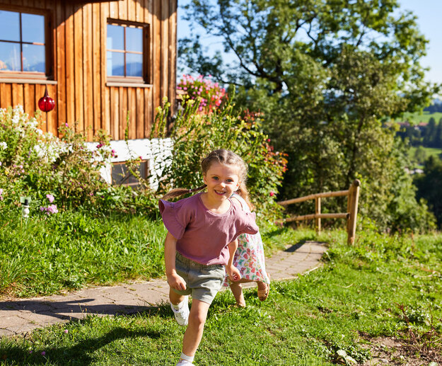 Das Bild zeigt ein junges Mädchen, das in einem pinken Hemd und grauen Shorts spazieren geht. Sie lächelt und scheint ihre Zeit im Freien zu genießen. Der Weg, auf dem sie spaziert, ist von einer Holzlattenwand an der einen Seite und einer Vielzahl von Blumen an der anderen Seite gesäumt. Das Haus im Hintergrund ist aus Holz, hat ein rotes Dach und sieht aus, als wäre es an einem klaren und blauen Himmel an einem sonnigen Tag. Die Gesamtszene wirkt friedlich und idyllisch, und der fröhliche Ausdruck des Mädchens verleiht dem Bild ein Gefühl von Wärme und Glück.