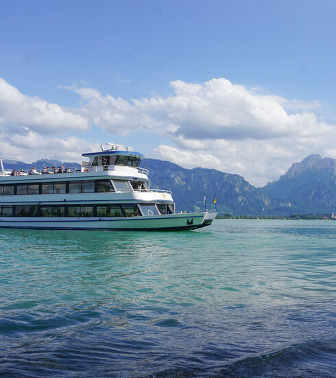 Das Bild fängt eine friedliche Szene eines weißen Schiffes mit drei Aussichtsdecks ein, das auf einem ruhigen blauen See – dem Forggensee - schwimmt. Das Schiff steht im Mittelpunkt des Bildes und wird von der unendlichen Weite des Forggensees umgeben. Der See selbst ist inmitten einer malerischen Kulisse von majestätischen Bergen eingebettet. Der klare und blaue Himmel ist vereinzelt von flauschigen, weißen Wolken durchzogen. 