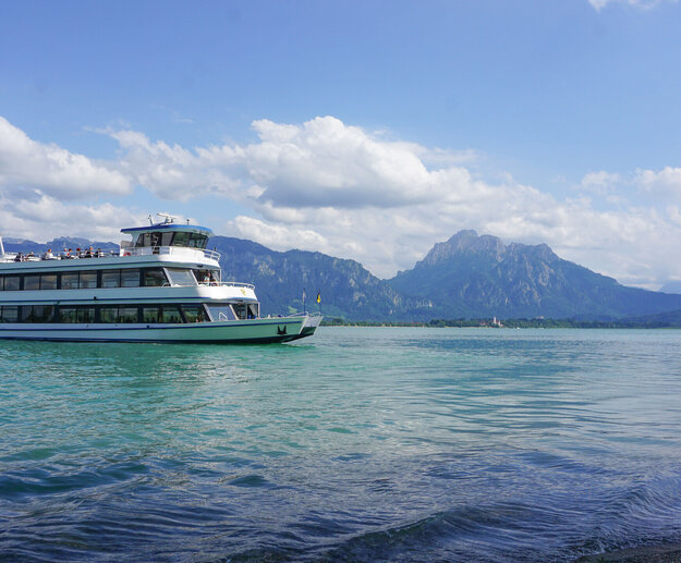 Das Bild fängt eine friedliche Szene eines weißen Schiffes mit drei Aussichtsdecks ein, das auf einem ruhigen blauen See – dem Forggensee - schwimmt. Das Schiff steht im Mittelpunkt des Bildes und wird von der unendlichen Weite des Forggensees umgeben. Der See selbst ist inmitten einer malerischen Kulisse von majestätischen Bergen eingebettet. Der klare und blaue Himmel ist vereinzelt von flauschigen, weißen Wolken durchzogen. 