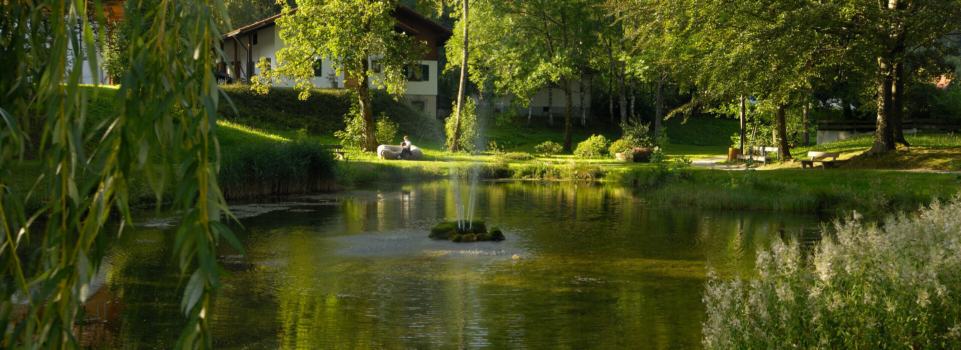Man blickt auf einen kleinen See, der inmitten eines Parks liegt. In der Mitte des Sees ragt eine Wasserfontäne in die Höhe. Der See ist von grünen Wiesen, hohen grünen Laubbäumen und Häuser umrahmt. Eine Person sitzt auf einer Bank und lies in einem Buch. Die Sonne scheint. Der Himmel ist blau. 