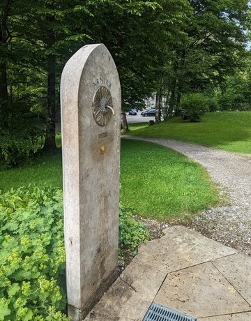  Das Bild zeigt ein schmales, längliches, steinernes Gebilde, das wie ein Denkmal aussieht. Die Struktur ist rechteckig geformt, mit einer gewölbten Oberseite. Es handelt sich um den Arnika Trinkbrunnen. Dieser befindet sich in einem parkähnlichen Areal, umgeben von üppigem grünem Gras und Bäumen. Der Brunnen ist mit einer Metallplatte verziert, die im oberen Drittel befestigt ist. Aus der Metallplatte ragt ein Wasserhahn. In die Metallplatte ist die Struktur einer Arnika-Blüte eingarbeitet.  Die Platte trägt die Inschrift "Arnika". Die gesamte Szene suggeriert eine friedliche und ruhige Umgebung. 