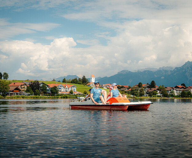 Das Bild zeigt eine vierköpfige Familie bestehend aus einem Mann, einer Frau einem Jungen und einem Mädchen, die auf einem Tretboot über einen See fahren. Sie tragen sportliche Freizeitkleidung und Sonnenbrillen. Am Ufer sind zahlreiche Häuser zu sehen. Im Hintergrund erheben sich die Berge. Der Himmel ist weiß-blau. Die Sonne scheint. 